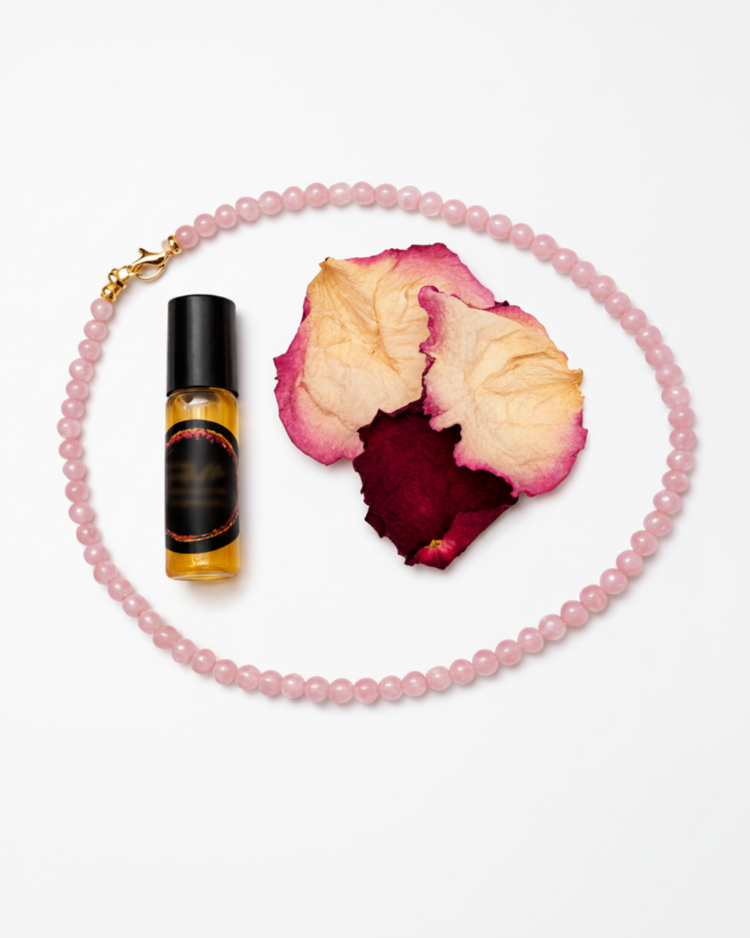 A rose quartz 4mm round beaded necklace with a gold clasp is arranged on a white background alongside dried rose petals and a small amber roller bottle with a black cap.