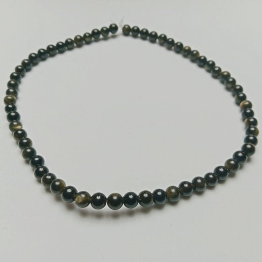 Macro studio photograph of polished Gold Sheen Obsidian beads catching direct light to display a rich gold flash across a high-gloss volcanic surface, styled at Belle Atelier.