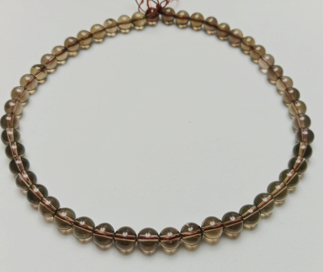 Macro studio photograph of translucent deep tea-brown and charcoal gray 6mm Smokey Quartz gemstone beads hand-assembled on a minimalist slate gray surface at Belle Atelier.