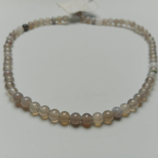 Macro studio photograph of a calibrated layered Gray Agate gemstone strand showing subtle white bands and cloudy gray textures, styled on a neutral stone slab at Belle Atelier.