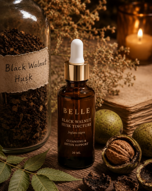 Belle Black Walnut Husk Tincture in an amber glass dropper bottle, styled on a rustic wooden table with whole and cracked walnuts, dried leaves, a jar of black walnut husk, and a burning candle in the background.