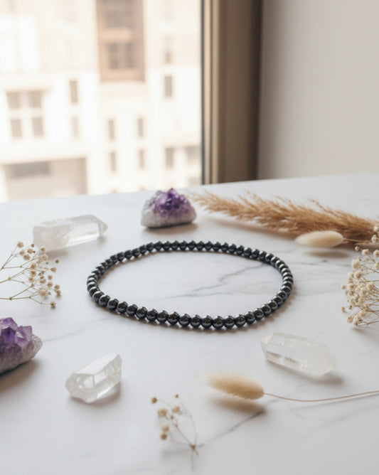 A high-end lifestyle flat lay displaying a polished 6mm hematite beaded necklace styled gracefully alongside raw quartz clusters and natural dried botanicals.
