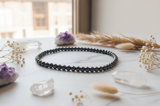 A high-end lifestyle flat lay displaying a polished 6mm hematite beaded necklace styled gracefully alongside raw quartz clusters and natural dried botanicals.
