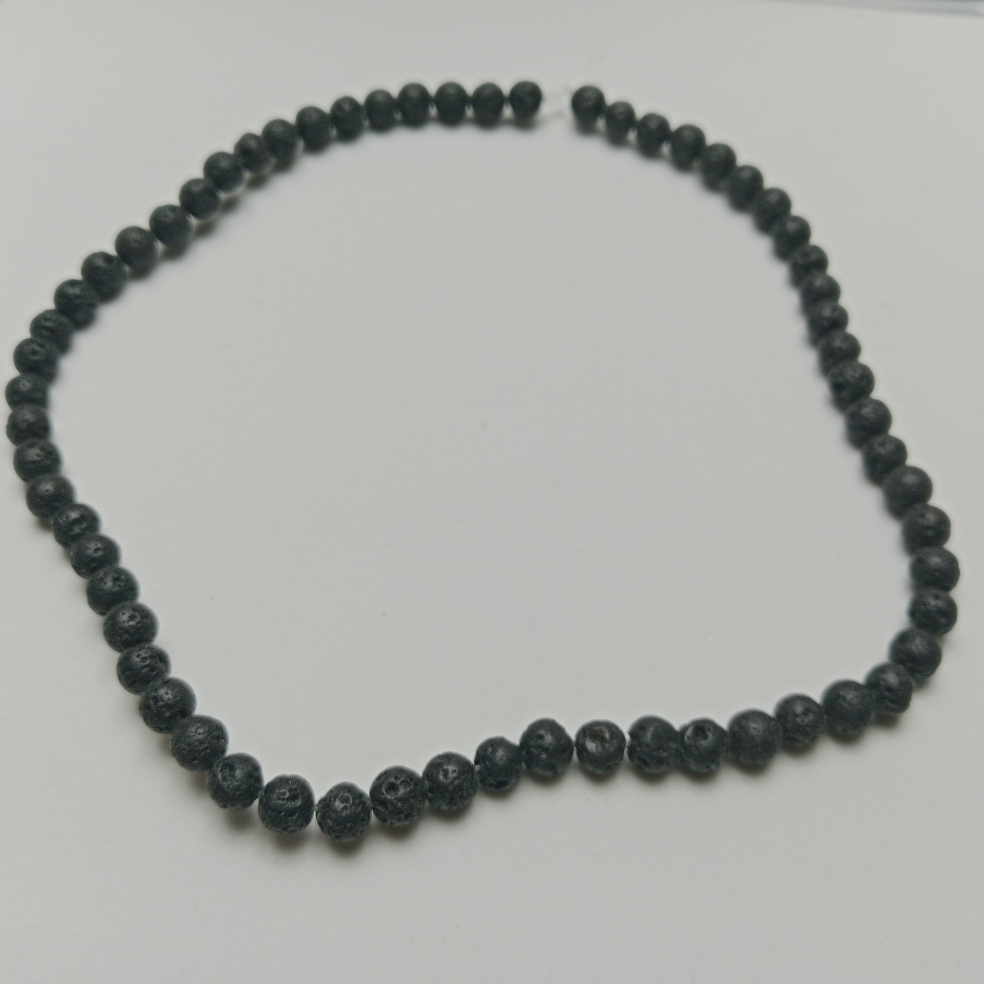 An overhead view of an unknotted 6mm black volcanic rock lava stone bead necklace strand arranged on a clean white surface.
