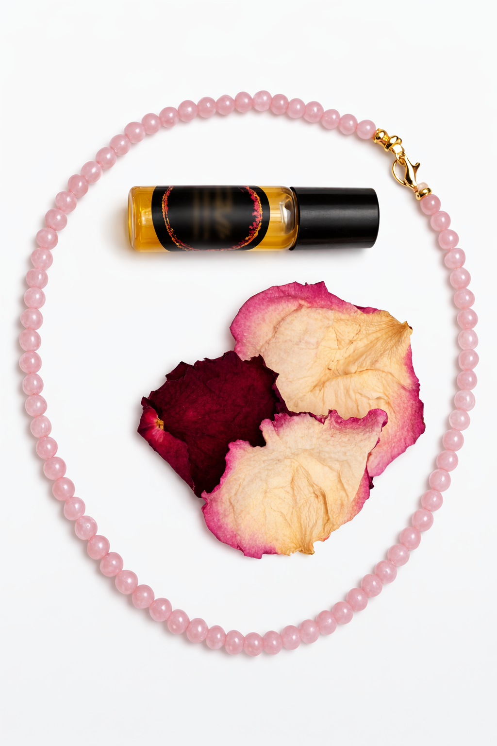 A rose quartz 4mm round beaded necklace with a gold clasp is arranged on a white background alongside dried rose petals and a small amber roller bottle with a black cap.