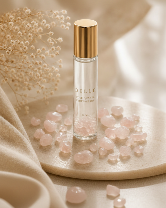 A clear glass bottle of BELLE Pure Quartz Perfume Oil with rose quartz and a shiny gold cap, styled on a beige stone tray with pink quartz crystals and dried flowers.