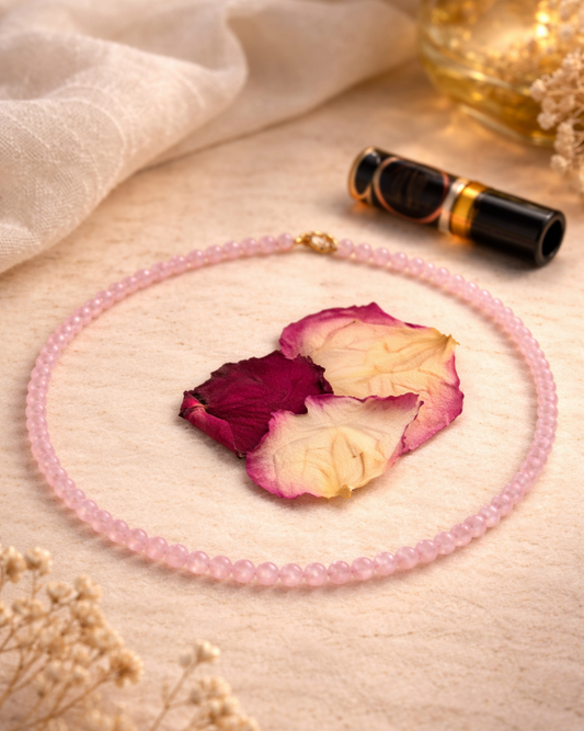 A pink beaded necklace with a small gold bead accent, arranged in a circle on a textured beige fabric alongside dried rose petals, a black and gold lip balm, and soft decorative flowers.