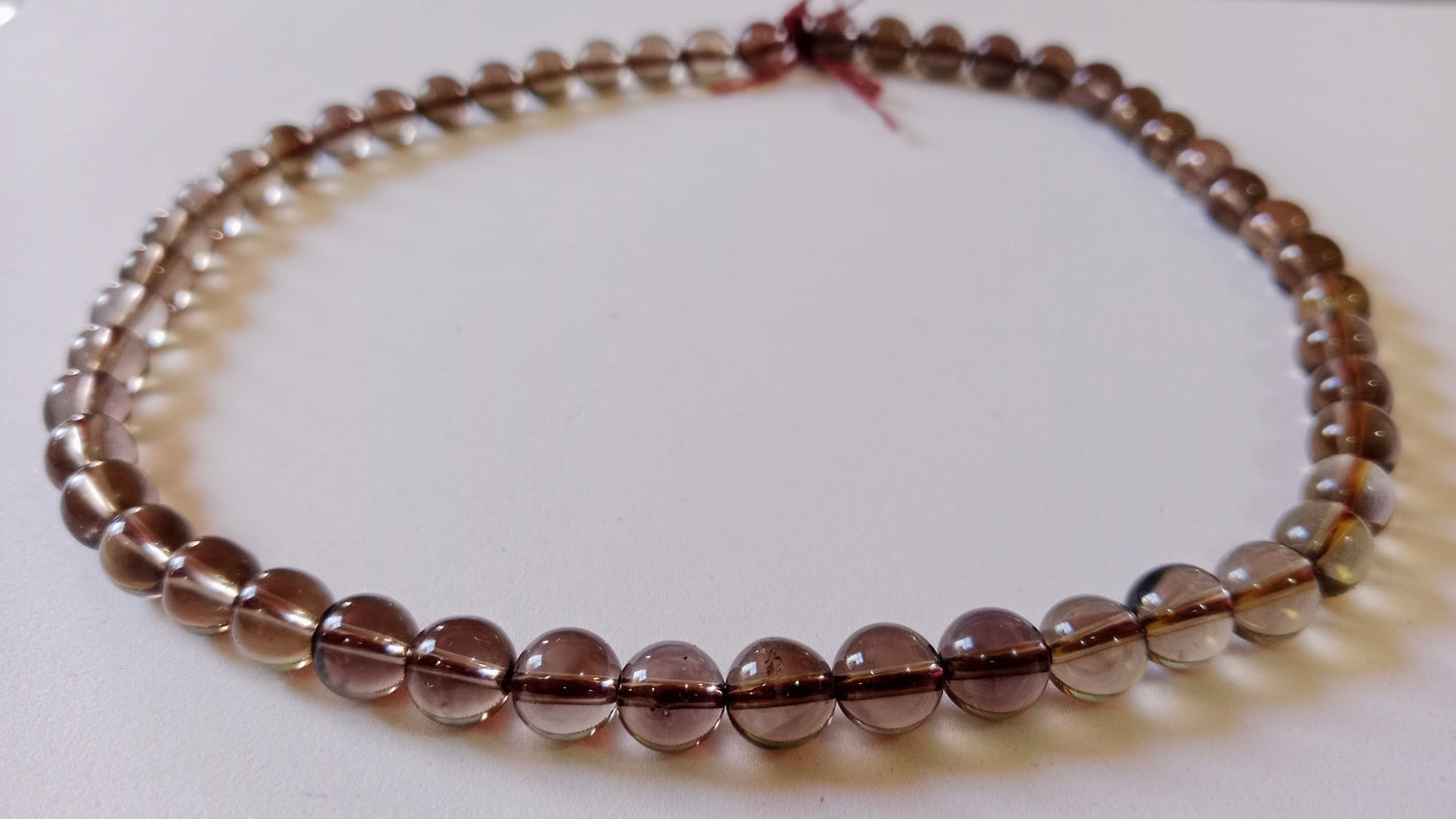 A high-end flat lay presentation of an unknotted smokey quartz bead necklace strand displaying translucent tea-brown gradients on a soft studio surface.
