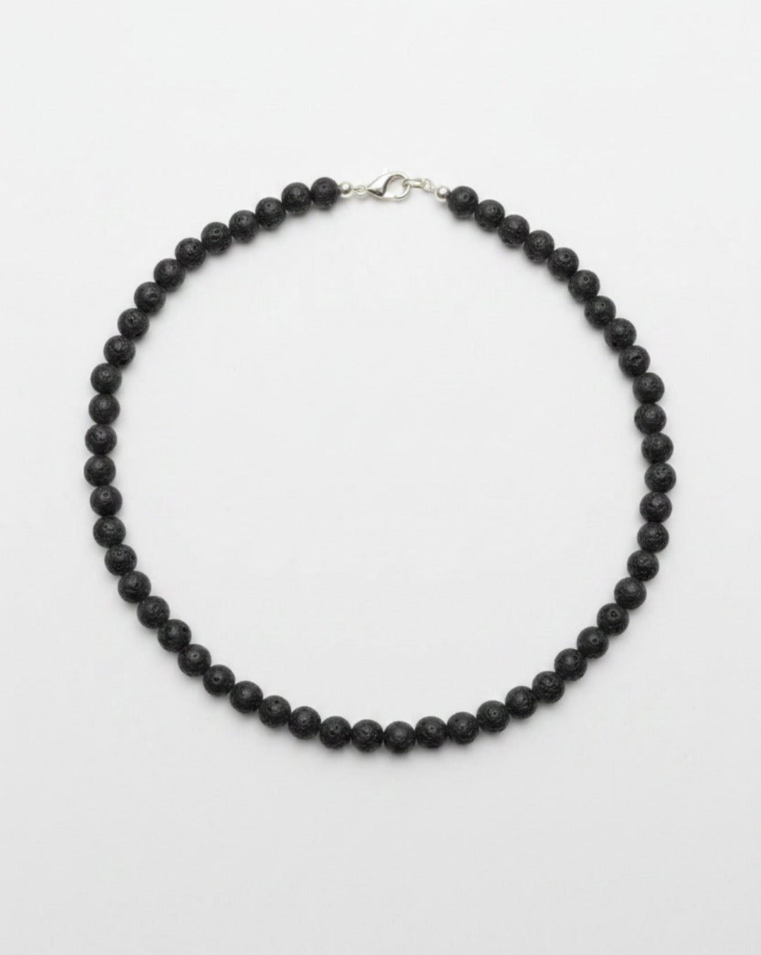 An overhead view of a raw natural 6mm black volcanic rock lava stone bead necklace strand arranged a clean white surface.
